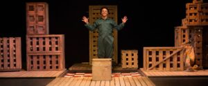 Image of performer from The Girl who Forgot to Sing Badly performance wearing a green jumpsuit standing beside wooden crates