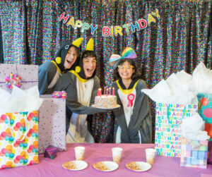 Three people dressed up as penguins at a birthday party. One of them is holding a birthday cake with candles.