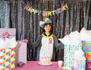 A female dressed as a penguin, standing in front of a birthday cake while surrounded by birthday presents.