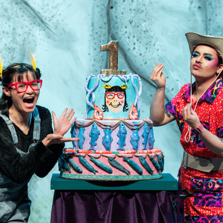 Two performers posing for a picture with a birthday cake; the person on the left is wearing red-rimmed eyeglasses, blue overalls and a black long-sleeved shirt. The person on the right is wearing a brightly-coloured safari outfit and a tan hat.