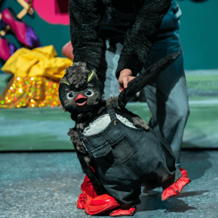 A penguin puppet dressed in overalls, being held by another performer