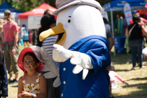 A smiling child stands next to a Seagull mascot wearing a blue shirt and a Port of Vancouver hat.