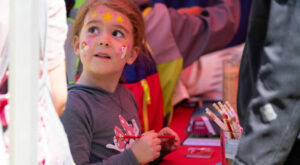 A child looks stands by an activity table, holding a pencil and looking to the left. Her forehead is painted with pink hearts and yellow stars; her left cheek has a rainbow and pink hearts; her right cheek has a unicorn and pink hearts.