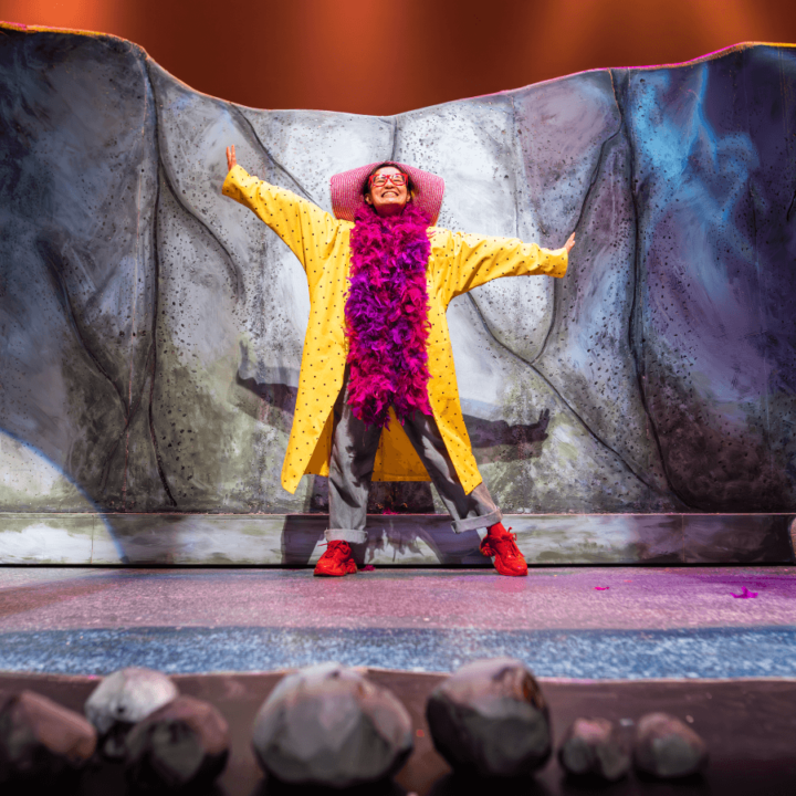 A performer in red-rimmed eyeglasses, a yellow coat, purple scarf and red shoes, stands on a stage.